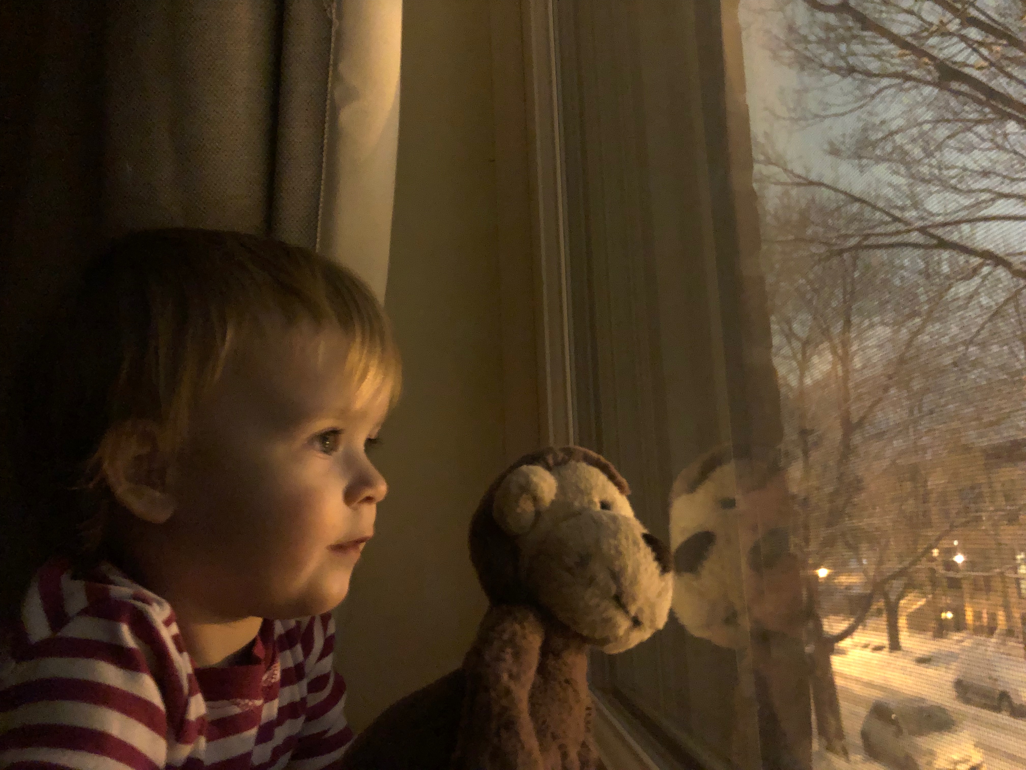 A young girl and a stuffed animal monkey looking out of a window together at a snowy street.